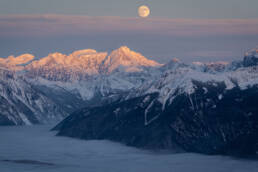 Snowy mountain range with a light blue sky and moon in the distance.